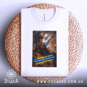 Футболка мама головнокомандувачка, білого кольору, складена на плоскій поверхні, принт з жінкою в халаті на коні, що вказує вперед, з яскравими кольорами та текстом, фон натуральний, з плетеним елементом.