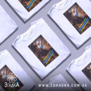 Футболка мама головнокомандувачка, білого кольору, зображена на моделі, яка стоїть на фоні світлого градієнта, принт з жінкою в халаті на коні, що вказує вперед, з яскравими кольорами та текстом.