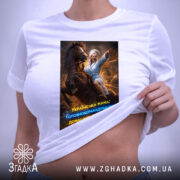 Футболка мама головнокомандувачка, білого кольору, зображена на плоскій поверхні, принт з жінкою в халаті на коні, що вказує вперед, з яскравими кольорами та текстом, на етикетці видно логотип бренду.