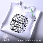Футболка мені завжди є що додати. Біла футболка, складена на столі, з чорним принтом, що містить текст. На футболці видно етикетку, а фон світло-сірий, що підкреслює чистоту дизайну.