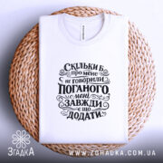 Футболка мені завжди є що додати. Біла футболка, складена на плетеному кошику, з чорним принтом, що містить текст. Фон світло-сірий, а текст на футболці чітко виділяється на білому матеріалі.