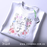 Футболка Мода це життя, біла футболка, знята зверху, на ній видно принт з квітами та написом 'Fashion is Life', на комірці є етикетка.