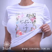 Футболка Мода це життя, біла футболка, знята зверху, на ній видно принт з квітами та написом 'Fashion is Life', на комірці є етикетка.