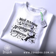 Футболка мої плани на вечір. Біла футболка, складена на плоскій поверхні, зображена зверху. На фоні світлий однотонний колір. Принт чорного кольору з написом "Мої плани на вечір: деградувати з комфортом" та ілюстрацією сидячої фігури з попкорном.