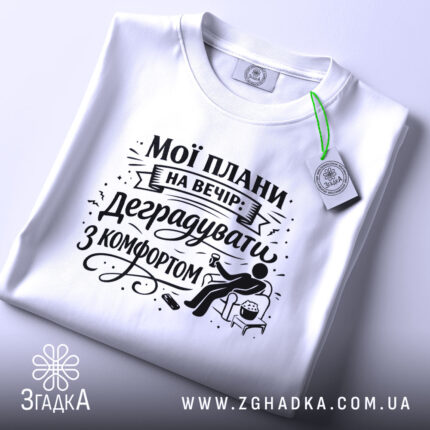 Футболка мої плани на вечір. Біла футболка, складена на плоскій поверхні, зображена зверху. На фоні світлий однотонний колір. Принт чорного кольору з написом "Мої плани на вечір: деградувати з комфортом" та ілюстрацією сидячої фігури з попкорном.