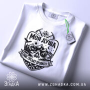 Футболка моя душа гори. Біла футболка, складена на поверхні, з чітким чорним принтом, що включає зображення гір, лісу та вогнища. На футболці видно етикетку з зеленим шнурком, що додає стильний акцент. Фон світлий, що підкреслює чистоту та простоту дизайну.