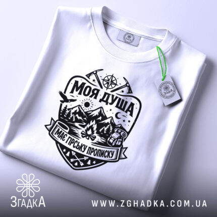Футболка моя душа гори. Біла футболка, складена на поверхні, з чітким чорним принтом, що включає зображення гір, лісу та вогнища. На футболці видно етикетку з зеленим шнурком, що додає стильний акцент. Фон світлий, що підкреслює чистоту та простоту дизайну.