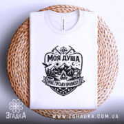Футболка моя душа гори. Біла футболка, складена на плетеній кошику, з чітким чорним принтом, що зображає гори, ліс та вогнище. Фон світлий, що підкреслює текстуру кошика, створюючи затишну атмосферу.