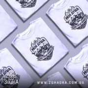 Футболка моя душа гори. Біла футболка з коротким рукавом, зображена на молодій людині, що позує з легким усмішкою. Принт на футболці чорного кольору, з елементами гір, сонця, лісу та рюкзака, що створює атмосферу пригод. Фон ніжно-фіолетовий, що підкреслює яскравість дизайну.