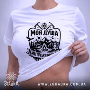 Футболка моя душа гори. Біла футболка, складена на поверхні, з чітким чорним принтом, що включає зображення гір, лісу та вогнища. На футболці видно етикетку з зеленим шнурком, що додає стильний акцент. Фон світлий, що підкреслює чистоту та простоту дизайну.