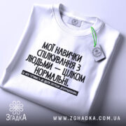 Футболка Моя терпимість, біла, лежить на плоскій поверхні, фон світло-сірий, принт чорного кольору з текстом на українській мові, етикетка з зеленим шнурком.