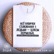 Футболка Моя терпимість, біла, лежить на плоскій поверхні з плетеним кошиком, фон білий, принт чорного кольору з текстом на українській мові, розташований по центру.