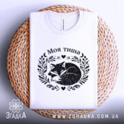 Футболка Моя тиша, біла, складена на плоскій поверхні, фон світло-сірий, принт з зображенням сплячого лиса в обрамленні рослинних мотивів і сердець.