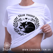Футболка Моя тиша, біла, на плоскій поверхні, з етикеткою, фон світло-сірий, принт з зображенням сплячого лиса в обрамленні рослинних мотивів і сердець.