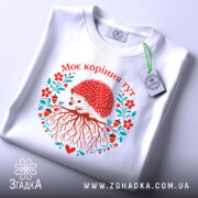 Футболка моє коріння тут, біла, зображена на плоскій поверхні, фон світло-сірий, принт з їжаком, червоними квітами та корінням, текст "Моє коріння тут" над принтом, з етикеткою.