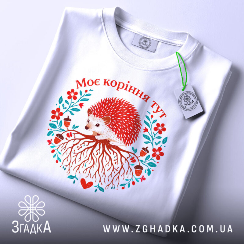 Футболка моє коріння тут, біла, зображена на плоскій поверхні, фон світло-сірий, принт з їжаком, червоними квітами та корінням, текст "Моє коріння тут" над принтом, з етикеткою.