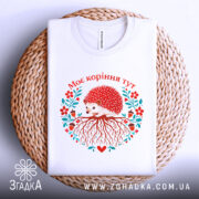 Футболка моє коріння тут, біла, зображена на плоскій поверхні, фон світло-сірий, принт з їжаком, червоними квітами та корінням, текст "Моє коріння тут" над принтом, на плетеній основі.