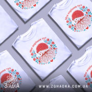 Футболка моє коріння тут, біла, зображена на моделі, фон світло-фіолетовий, принт з їжаком, червоними квітами та корінням, текст "Моє коріння тут" над принтом.