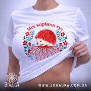 Футболка моє коріння тут, біла, зображена на плоскій поверхні, фон світло-сірий, принт з їжаком, червоними квітами та корінням, текст "Моє коріння тут" над принтом, з етикеткою.