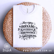 Футболка Моє куку поїхало, біла, на плоскій поверхні, фон світло-сірий, принт чорного кольору з текстом, футболка розташована на плетеній основі.