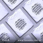 Футболка Моє куку поїхало, біла, на моделі з коротким рукавом, фон світло-фіолетовий, принт чорного кольору з текстом і графічними елементами.