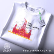 Футболка москва у вогні, складена футболка на білому фоні, ракурс зверху, принт з червоним і жовтим зображенням Кремля з написом 'I'M A FIRESTARTER' та 'MOSCOW ON FIRE', етикетка на футболці.