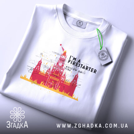 Футболка москва у вогні, складена футболка на білому фоні, ракурс зверху, принт з червоним і жовтим зображенням Кремля з написом 'I'M A FIRESTARTER' та 'MOSCOW ON FIRE', етикетка на футболці.