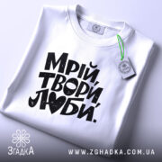 Футболка Мрій Твори Люби, біла, на плоскій поверхні, з ракурсом зверху, фон світло-сірий, принт чорного кольору з написом 'Мрій, твори, люби.' без додаткових елементів.