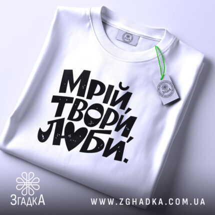 Футболка Мрій Твори Люби, біла, на плоскій поверхні, з ракурсом зверху, фон світло-сірий, принт чорного кольору з написом 'Мрій, твори, люби.' без додаткових елементів.