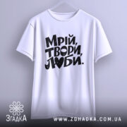 Футболка Мрій Твори Люби, біла, підвішена на вішалці, з ракурсом спереду, фон світло-сірий, принт чорного кольору з написом 'Мрій, твори, люби.' в стильному виконанні.