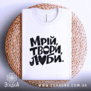 Футболка Мрій Твори Люби, біла, на плоскій поверхні, з ракурсом зверху, фон світло-сірий, принт чорного кольору з написом 'Мрій, твори, люби.' на фоні плетеного кошика.