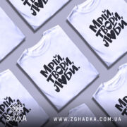 Футболка Мрій Твори Люби, біла, на моделі, з легким ракурсом спереду, фон світло-фіолетовий, принт чорного кольору з написом 'Мрій, твори, люби.' у креативному стилі.