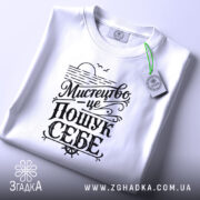 Футболка мистецтво це пошук себе. Біла футболка на плоскій поверхні, зображена зверху, фон світло-сірий, принт чорного кольору з написом 'Мистецтво це пошук себе', на футболці є етикетка.