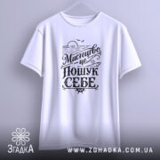 Футболка мистецтво це пошук себе. Біла футболка, підвішена на вішалці, фон світло-сірий, принт чорного кольору з написом 'Мистецтво це пошук себе', видно округлі краї рукавів.