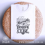 Футболка мистецтво це пошук себе. Біла футболка на плоскій поверхні, зображена зверху, фон світло-сірий, принт чорного кольору з написом 'Мистецтво це пошук себе', футболка лежить на плетеній основі.