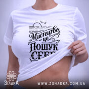 Футболка мистецтво це пошук себе. Біла футболка на плоскій поверхні, зображена зверху, фон світло-сірий, принт чорного кольору з написом 'Мистецтво це пошук себе', на футболці є етикетка.