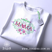 Футболка найкраща у світі мама, біла, складена на поверхні, фон світлий, принт з квітами та написом 'НАЙКРАЩА у світі МАМА', з етикеткою на комірці.