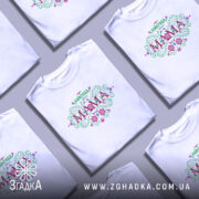 Футболка найкраща у світі мама, біла, зображена на моделі з легким ракурсом, фон ніжно-фіолетовий, принт з квітковими елементами та написом 'НАЙКРАЩА у світі МАМА'.