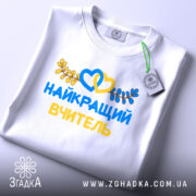 Футболка найкращого вчителя випускників, біла, складена на поверхні, фон світлий, принт з жовтими та блакитними елементами, текст "НАЙКРАЩИЙ ВЧИТЕЛЬ" та декоративні листя, на комірці видна етикетка.