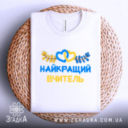 Футболка найкращого вчителя випускників, біла, складена на плетеній основі, фон світлий, принт з жовтими та блакитними елементами, текст "НАЙКРАЩИЙ ВЧИТЕЛЬ" та декоративні листя.