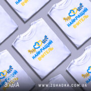 Футболка найкращого вчителя випускників, біла, зображена на моделі з легким макіяжем, фон ніжно-фіолетовий, принт з жовтими та блакитними елементами, текст "НАЙКРАЩИЙ ВЧИТЕЛЬ" та декоративні листя.