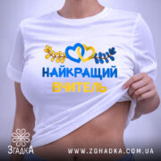 Футболка найкращого вчителя випускників, біла, складена на поверхні, фон світлий, принт з жовтими та блакитними елементами, текст "НАЙКРАЩИЙ ВЧИТЕЛЬ" та декоративні листя, на комірці видна етикетка.