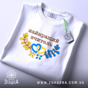 Футболка найкращому вчителю, біла, на столі, фон світло-сірий, принт з написом 'найкращий вчитель' коричневого кольору, оформлений жовтими та блакитними елементами у вигляді сердець та гілок, етикетка на комірці.