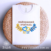 Футболка найкращому вчителю, біла, на плетеній основі, фон світло-сірий, принт з написом 'найкращий вчитель' коричневого кольору, оформлений жовтими та блакитними елементами у вигляді сердець та гілок.