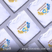 Футболка найкращому вчителю, біла, на моделі, фон світло-сірий, принт з написом 'найкращий вчитель' коричневого кольору та елементами жовтого і блакитного, оформлена у вигляді сердець та гілок.