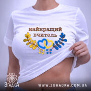 Футболка найкращому вчителю, біла, на столі, фон світло-сірий, принт з написом 'найкращий вчитель' коричневого кольору, оформлений жовтими та блакитними елементами у вигляді сердець та гілок, етикетка на комірці.