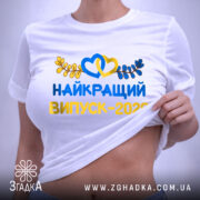 Футболка найкращий випускний 2026, біла, на столі, з видимим етикеткою, фон світло-сірий, принт яскравий, з сердечками та листям, напис великими літерами.