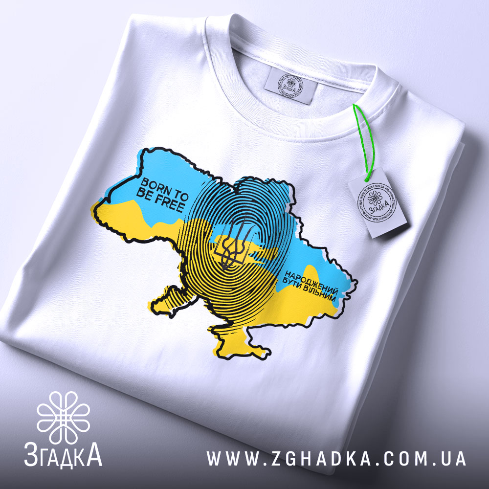 Футболка народжений вільним, біла, на столі, з видимим принтом карти України в жовто-блакитних кольорах, текстом 'BORN TO BE FREE' і 'НАРОДЖЕНИЙ БУТИ ВІЛЬНИМ', етикетка на зеленому шнурку.