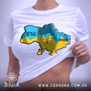 Футболка народжений вільним, біла, на столі, з видимим принтом карти України в жовто-блакитних кольорах, текстом 'BORN TO BE FREE' і 'НАРОДЖЕНИЙ БУТИ ВІЛЬНИМ', етикетка на зеленому шнурку.