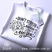 Футболка Не стресуй мені мозок. Біла футболка, складена на поверхні, з видимим принтом чорного кольору. Текст 'DON'T STRESS мені мозок' та малюнок мозку. Фон світло-сірий, з елементами освітлення.